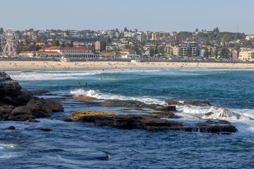 Obraz premium Waves washing up on rocks in the foreground, before iconic Bondi Beach and Bondi Pavilion, in Sydney, Australia. People enjoying the sand and water.