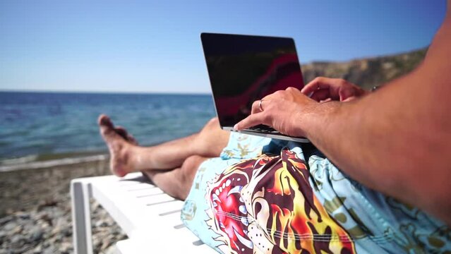 Man laptop sea. Working remotely on seashore. Happy successful man, male freelancer working on laptop on beach, relieves stress from work to restore life balance. Freelance, remote work on vacation