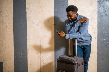 Happy man with suitcase using phone while leaning against a  wall in  passage of the railway station. Copy space on image for your advert or text.