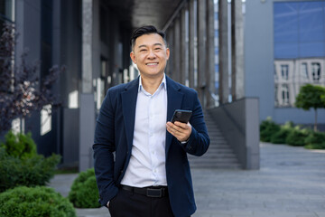 Portrait of Asian successful young Asian man businessman, lawyer, politician in suit standing near office building holding phone and hand in pocket, smiling looking at camera
