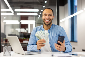 Portrait of a happy young hispanic man sitting in the office at the table, holding cash money and a...