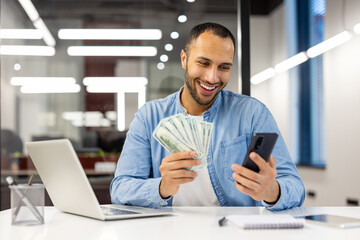 Smiling hispanic young man sitting in a formal office at a table, holding cash money banknotes in his hands, using a mobile phone