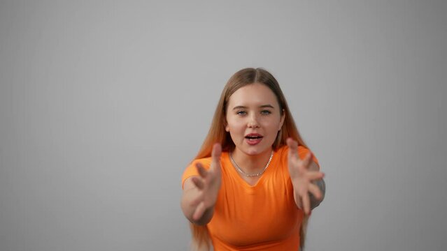 Portrait of young stressed shouting girl. Amazed excited young woman in orange t-shirt screaming raising hands opening mouth closing eyes in studio indoors. Surprise, amazement, panic concept.
