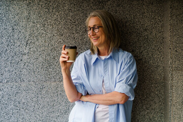 A woman wearing glasses stands holding a cup of coffee.