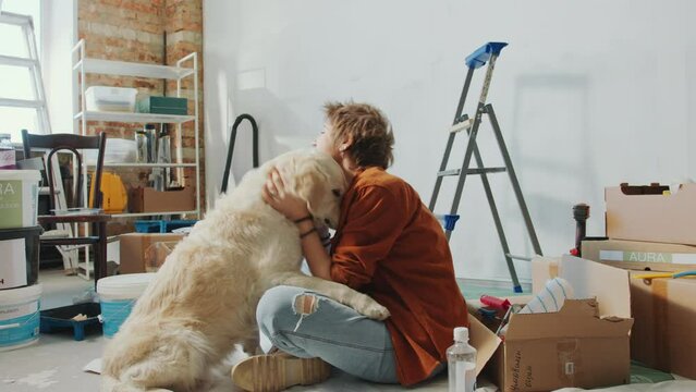 Young happy woman sitting on the floor among boxes and paint buckets, petting lovely golden retriever dog and smiling when renovating home by herself