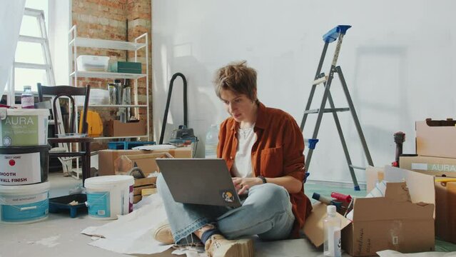 Young woman sitting cross legged on the floor among paint buckets and boxes, searching home renovation ideas on the Internet and looking around. Zoom Shot