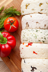 Closeup of several varieties of homemade cheese with different fillings with paprika, dill, olives, walnuts and cookies on a wooden board with fresh dill, tomatoes and bell pepper. - 765694079