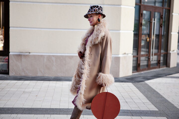 Fashionable woman walking in the city, dressed in a long beige coat, Cossack boots, hat with animal...