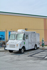 A white food truck, somewhat worn, is parked next to a yellow building with large windows; seems to be ready to start selling food.