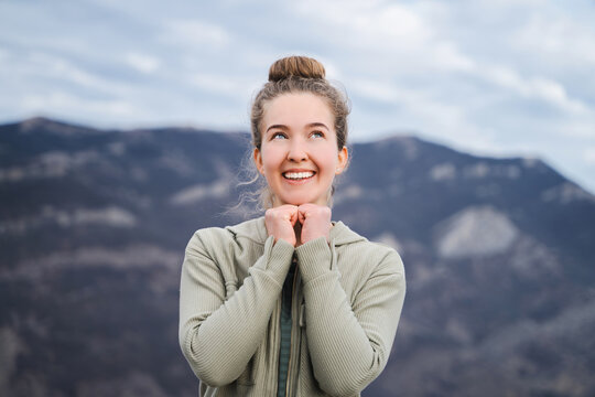 Young cheerful thoughtful woman with curly hair toothy smiling  leaned on hands look empty space in the mountains, portrait of a good mood dreamy girl in casual wear arms cheeks looking up