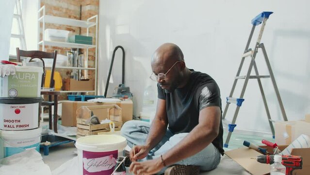 Young African American man sitting on the floor in living room with home improvement supplies and mixing paint colors in cup before painting walls
