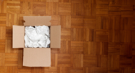 Cardboard box filled with shipping packaging paper on a wood floor, seen directly from above in...
