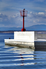 Red metal lighthouse on white stone pier with reflection on sea surface