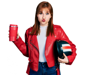 Redhead young woman holding motorcycle helmet and take away coffee puffing cheeks with funny face. mouth inflated with air, catching air.