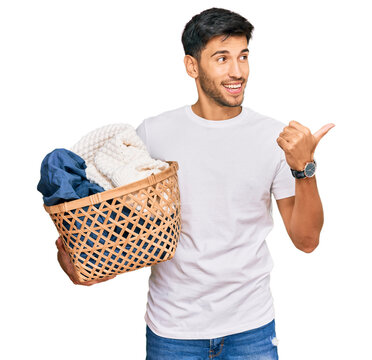 Young handsome man holding laundry basket pointing thumb up to the side smiling happy with open mouth