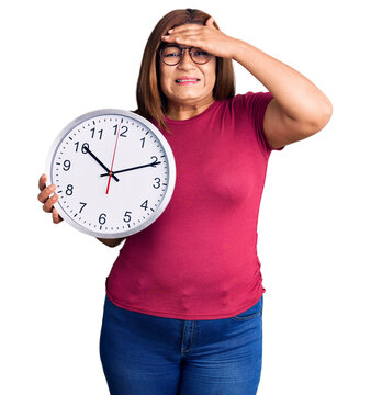 Middle age latin woman holding big clock stressed and frustrated with hand on head, surprised and angry face