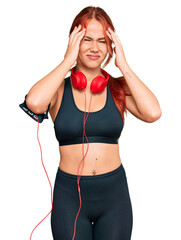 Young redhead woman wearing gym clothes and using headphones suffering from headache desperate and stressed because pain and migraine. hands on head. - obrazy, fototapety, plakaty