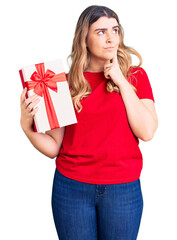 Young caucasian woman holding gift serious face thinking about question with hand on chin, thoughtful about confusing idea