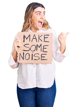 Young caucasian woman holding make some noise banner pointing thumb up to the side smiling happy with open mouth