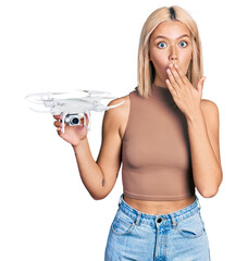Beautiful young blonde woman holding drone covering mouth with hand, shocked and afraid for...