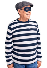 Senior handsome man wearing burglar mask and t-shirt looking away to side with smile on face,...