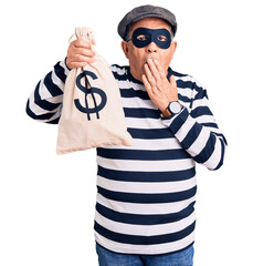 Senior handsome man wearing burglar mask holding money bag covering mouth with hand, shocked and...