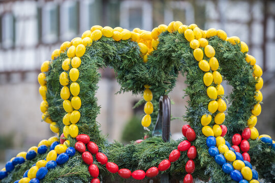 Osterbrunnen mit gelben, roten und blauen Ostereiern aus Kunststoff vor historischem Geb&auml;ude Pfarrhaus in Neckartenzlingen