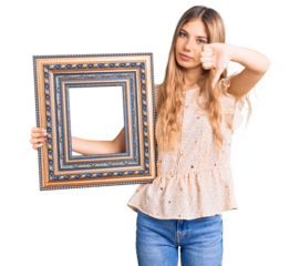 Beautiful caucasian woman with blonde hair holding empty frame with angry face, negative sign showing dislike with thumbs down, rejection concept