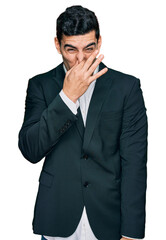 Handsome hispanic man wearing business clothes smelling something stinky and disgusting, intolerable smell, holding breath with fingers on nose. bad smell