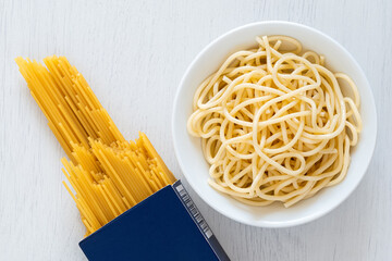 Cooked spaghetti in a white ceramic bowl next to uncooked spaghetti in a blue box on white painted wood. - 765782422
