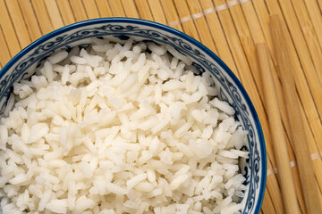 Detail of cooked white rice in an asian white and blue ceramic bowl next to chopsticks isolated on light bamboo matt. Top view.