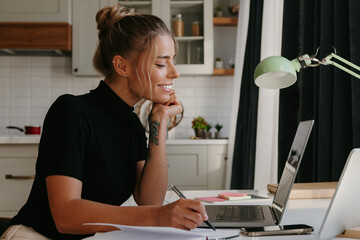 Smiling young woman making notes and looking at the laptop while working at home - 765784095