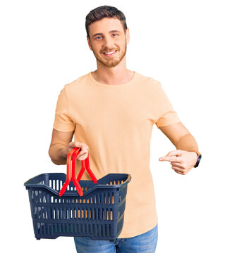 Handsome young man with bear holding supermarket shopping basket smiling happy pointing with hand and finger