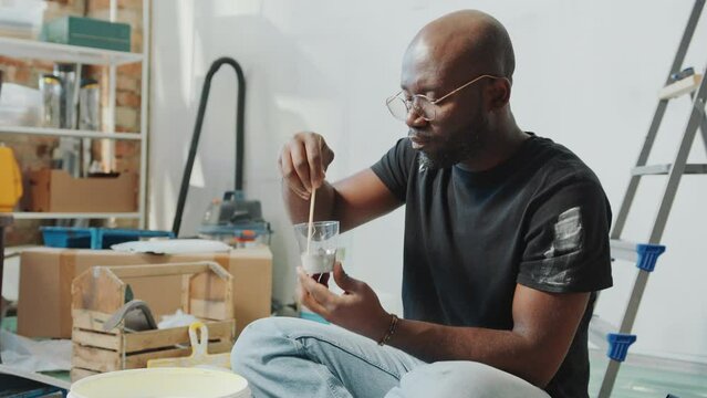 Black man in stained t-shirt sitting on the floor in living room, mixing paint in cup and examining its color when doing DIY home renovation
