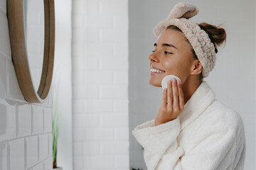 Beautiful young woman in bathrobe cleaning face with cotton pad while standing in bathroom - 765785484