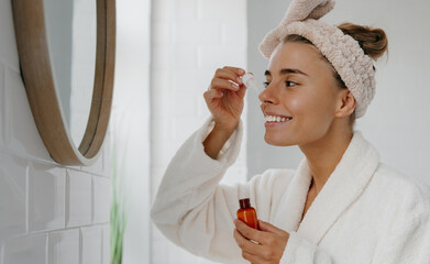 Beautiful young woman in bathrobe applying cosmetic serum on face while standing in bathroom