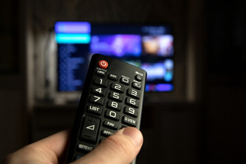 A person switches channels on a TV using a remote control. In the hand of a person, a TV remote control, a close-up photo, against the background of a blurred TV