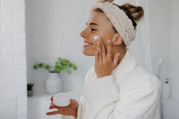 Happy young woman in bathrobe applying face cream while standing in bathroom - 765794234