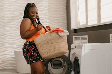 Happy plus size African woman carrying basket with clothes while standing near the washing machine in bathroom