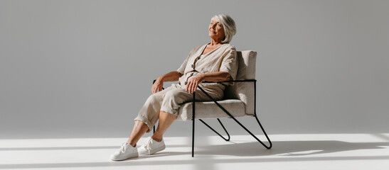 Relaxed gray hair senior woman sitting in comfortable chair and looking calm against background - 765796295