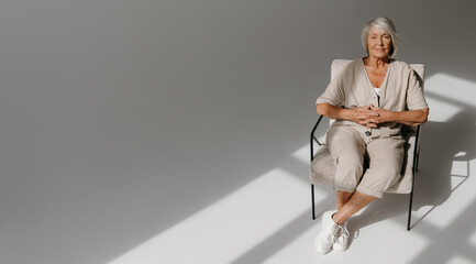 Top view of calm gray hair senior woman relaxing in comfortable chair in sunny studio