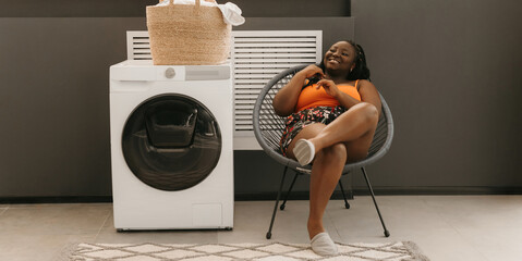 Happy plus size African woman waiting for clean clothes while sitting in chair near the washing machine in laundry - 765801245