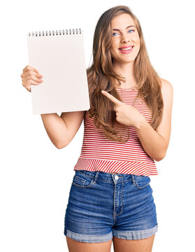 Beautiful caucasian young woman holding notebook smiling happy pointing with hand and finger