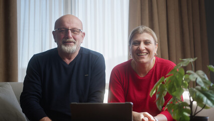 Elderly couple sitting on the couch looking into the camera, using webcam to talking with...