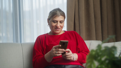 Middle aged mature woman sit on couch holding smartphone looking at screen using social media apps, enjoying free time. Older people and technology concept	
