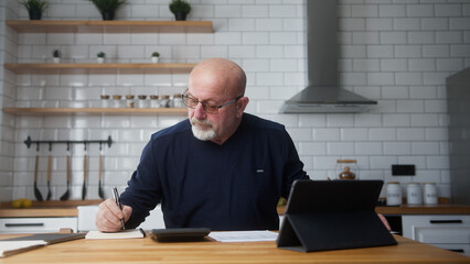 Old man sit in the kitchen using tablet device, study financial papers using calculator count...