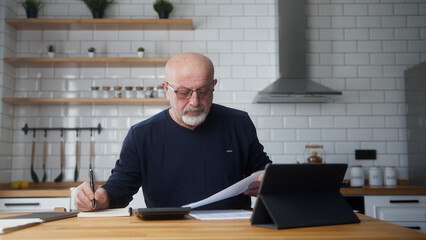 Elderly man sit in the kitchen using tablet device, study financial papers using calculator count...