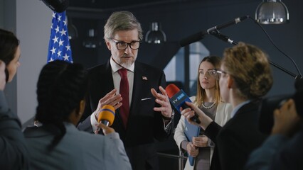 Serious male American politician talks with journalists, answers questions and gives interview for media and television news in government building. United States congressman during press conference.