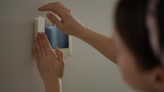 Close-up. A woman carefully covers the display of a video intercom with masking tape before painting the walls, standing in the hallway of her apartment. Paint a room DIY