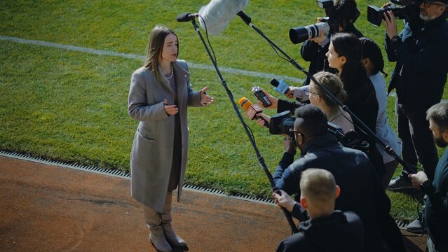 Female director of football team answering press questions and giving interview near soccer field before important match. Investor or businesswoman crowded by news journalists. Inspirational speech.
